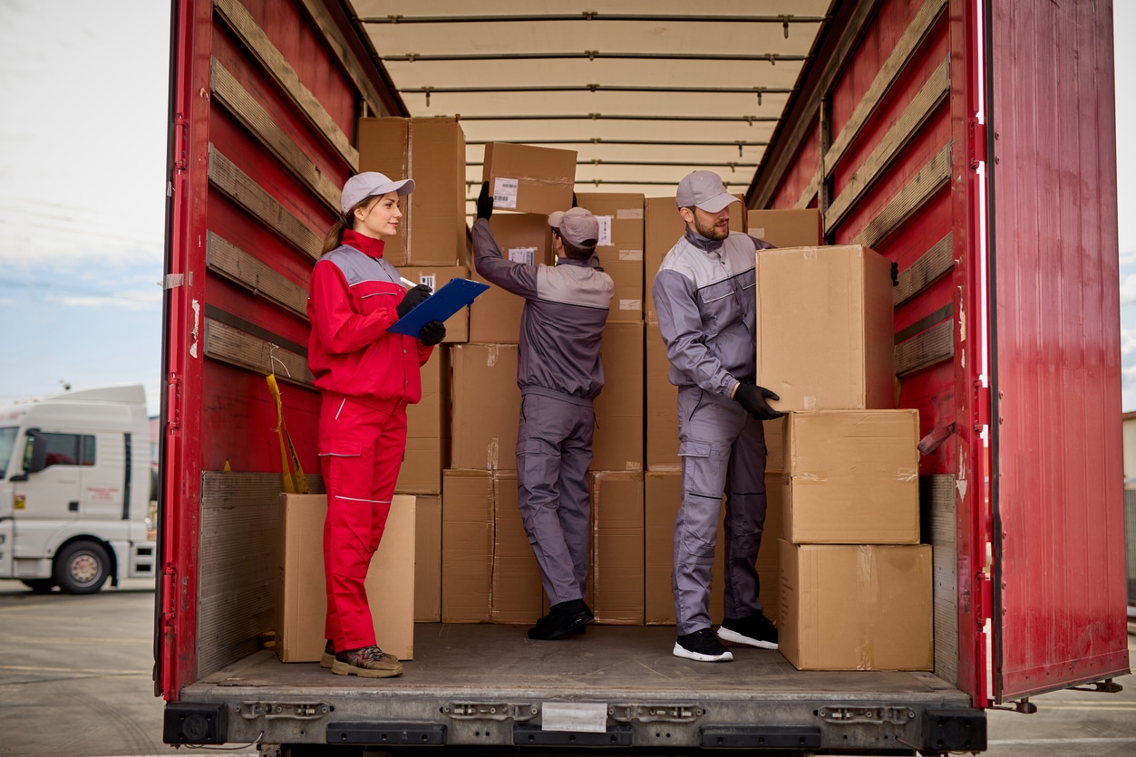 A woman with a clipboard stands in the back of a semi-truck, while two men move boxes.