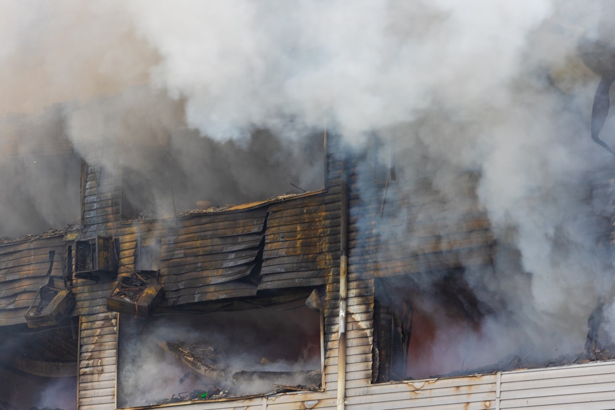 Thick smoke pours out of a building engulfed in fire while the siding is warped and discolored.